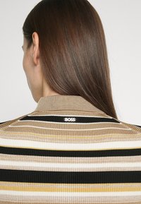 Personne aux longs cheveux bruns portant un pull côtelé beige, noir, blanc et jaune à rayures avec le logo "BOSS" sur le col arrière.