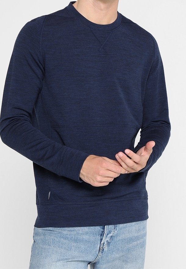 Homme portant un sweat-shirt bleu marine à manches longues et un jean bleu clair, ajustant le poignet gauche avec les mains.