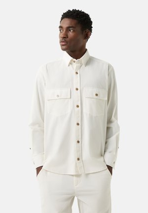 Mand iført hvid skjorte med lange ærmer og knapper, med brystlommer, samt hvide bukser, kigger mod venstre mod en ensfarvet baggrund.