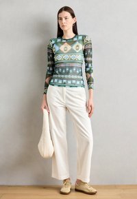 Top in rete a maniche lunghe con motivi geometrici verdi e blu, abbinato a pantaloni bianchi a gamba larga e sneaker beige, mentre tiene una borsa morbida beige.
