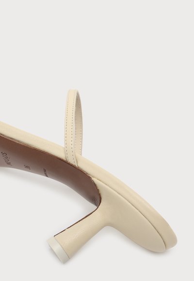 Gros plan d'une sandale à talon beige avec une fine bride à l'arrière et une semelle intérieure marron sur fond blanc uni.