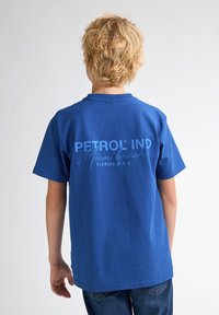 Modré bavlněné tričko s nápisem "PETROL IND Miami Beach, Florida, U.S.A." ve světle modré barvě na zadní straně. Krátké rukávy a kulatý výstřih.
