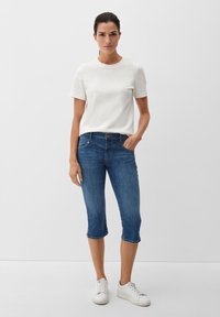 Weißes Kurzarmhemd, blaue Jeans-Capri im schlanken Design und weiße Sneakers, stehend vor einem schlichten Hintergrund.