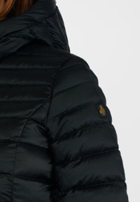Giacca a vento nera imbottita con tessuto trapuntato e cuciture orizzontali, colletto alto e una piccola etichetta con logo sulla manica.