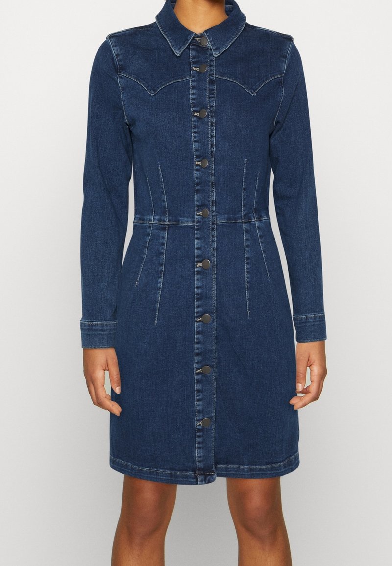 Personne portant une robe en jean bleu foncé à manches longues et longueur genou, avec des boutons sur le devant et un col pointu, debout devant un fond uni.