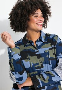 Camisa con patrón de camuflaje en verde, azul y negro. Presenta un cuello clásico, botones a presión y detalles de lentejuelas en las mangas.