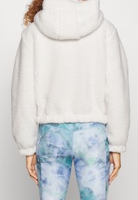 Chaqueta corta de forro polar blanca con cuello alto y superficie texturizada, combinada con leggings de teñido anudado en azul y verde.