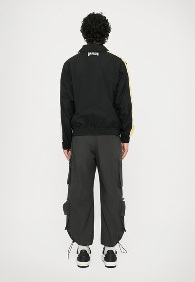 Giacca a vento nera con strisce gialle sui lati, polsini elasticizzati e un logo sulla schiena, abbinata a pantaloni cargo grigi larghi e scarpe nere.