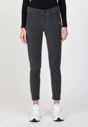 Osoba ubrana w dopasowane ciemnoszare sztruksowe spodnie, czarną bluzkę z długim rękawem oraz białe sportowe buty, stojąca na jednolitym tle.