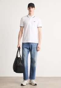 Biely polo trik s čiernym lemovaním, modré džínsy, čierna kabelka a šedé tenisky. Neformálny outfit na stojacom mužskom figure na neutrálnom pozadí.