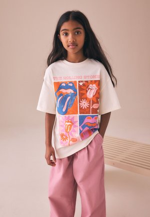 Młoda dziewczyna z długimi ciemnymi włosami, ubrana w białą koszulkę Rolling Stones i luźne różowe spodnie, stoi przy drewnianej ławce.
