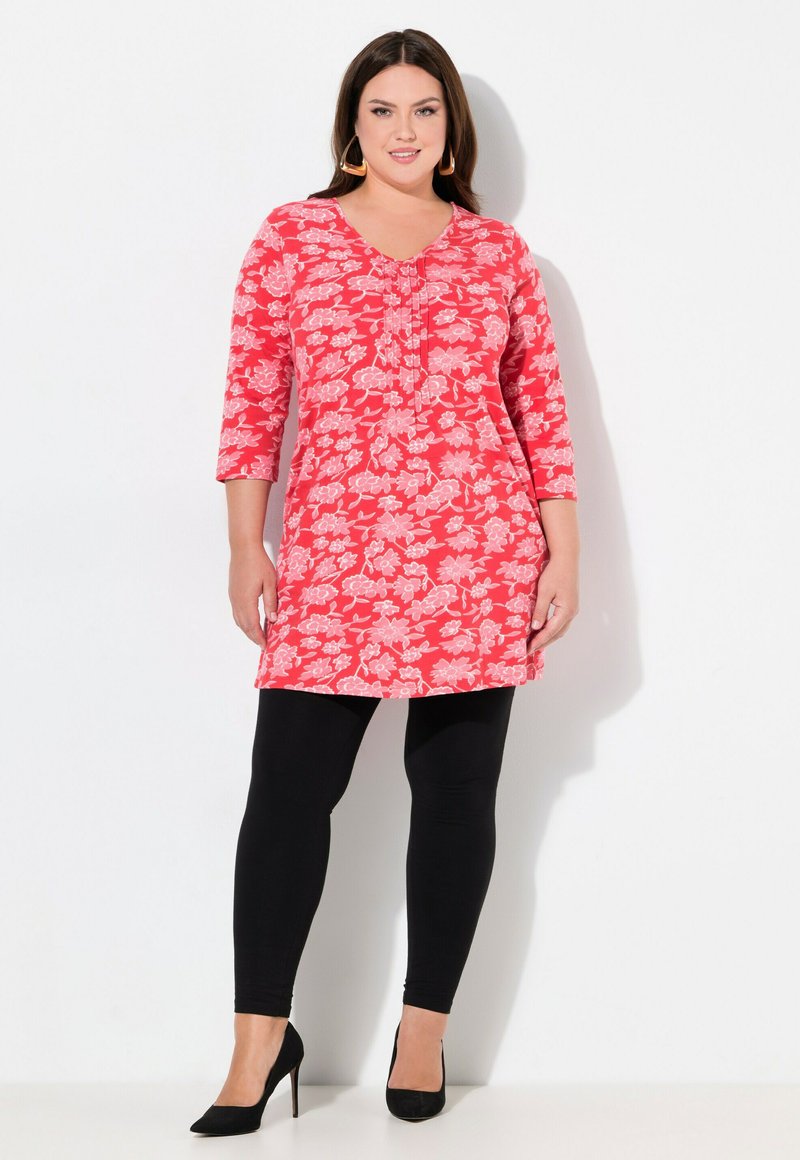 Kwiatowa tunika w jaskrawym czerwonym kolorze z białymi wzorami kwiatowymi, z dekoltem w kształcie litery V, rękawami o długości trzech czwartych i plisowanym przodem. Połączona z czarnymi legginsami.