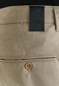 Beżowe spodnie z teksturowanej tkaniny, z czarną skórzaną metką na pasku i brązowym guzikiem na tylnej kieszeni.