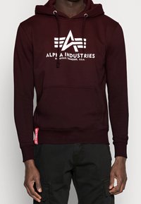 Uomo che indossa un hoodie Alpha Industries di un rosso scuro con logo bianco e pantaloni cargo neri, in piedi contro uno sfondo chiaro e semplice.