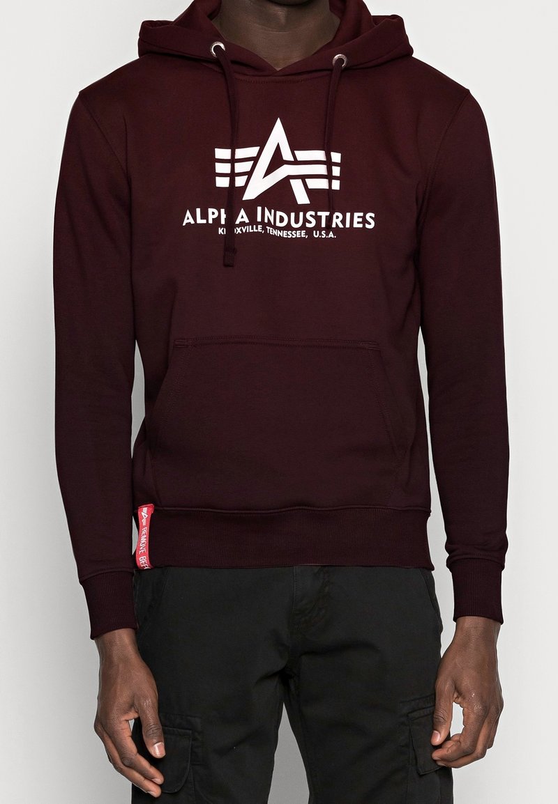 Uomo che indossa un hoodie Alpha Industries di un rosso scuro con logo bianco e pantaloni cargo neri, in piedi contro uno sfondo chiaro e semplice.