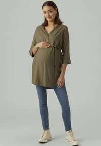 Túnica longa verde-oliva com uma pala de botões, amarrada abaixo do busto, combinada com jeans skinny azuis e sapatilhas de cano alto bege.