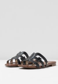 Braune flache Sandalen mit offener Zehenpartie und schwarzen Leder-Schlitzriemen, auf einer weißen Oberfläche mit einem schlichten Hintergrund präsentiert.