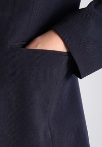 Marineblazer aus strukturiertem Stoff, mit einer versteckten Seitentasche und einem taillierten Slim-Fit-Design. Nahaufnahme des Ärmels und der Taschedetails.