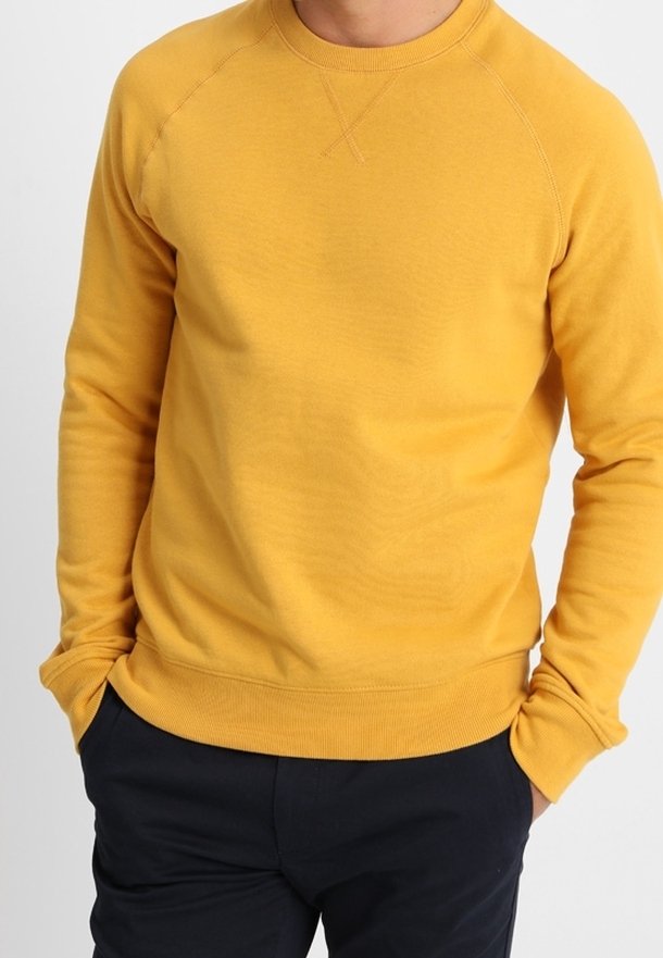 Homme portant un sweat-shirt jaune moutarde à encolure ronde avec manches longues et pantalon foncé, debout les mains dans les poches.