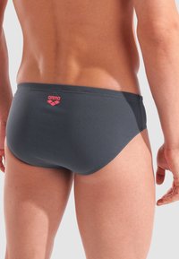 La parte bassa della schiena e le gambe superiori di un uomo che indossa slip da bagno Arena grigio scuro con logo rosso, su uno sfondo chiaro e semplice.