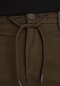 Brune bukser med struktureret overflade, udstyret med elastisk talje og lukning med metalknap. Der er detaljer med matchende tråd og aglet.