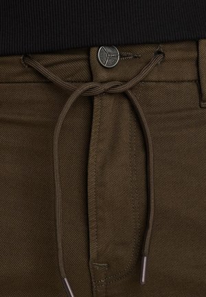 Smeđe hlače s teksturiranim finišem, s pojasom na vezanje i metalnim dugmetom za zatvaranje. Akcenti uključuju usklađene konce i detalje od agleta.