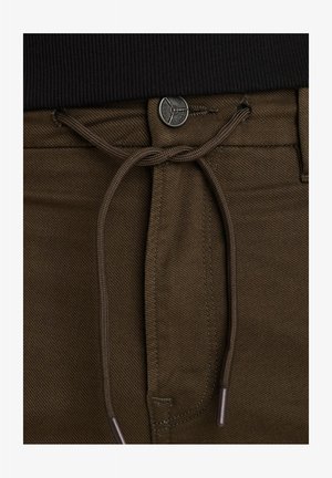 Smeđe hlače s teksturiranim finišem, s pojasom na vezanje i metalnim dugmetom za zatvaranje. Akcenti uključuju usklađene konce i detalje od agleta.