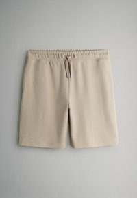 Beżowe szorty z prążkowanej tkaniny z elastycznym paskiem w talii i przednim ściągaczem na jednolitym jasnoszarym tle.