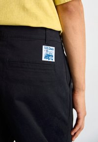 Svarta byxor i tyg har en bakficka och en etikett med texten "TUFF-ONES by Champion" med en blå grafisk design.