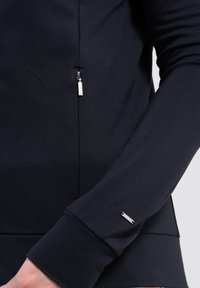 Haut à manches longues bleu marine avec une texture lisse, comportant une poche zippée et un accent de logo argenté sur le poignet. Design simple et ajusté.