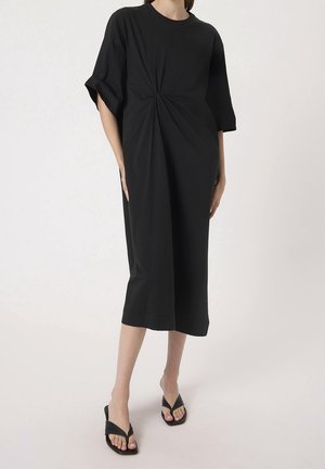 Femme portant une robe midi noire avec un détail de nœud central, associée à des sandales à talons noires, debout devant un fond clair uni.