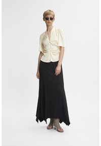 Blusa crema con maniche a sbuffo e scollo a V, abbinata a una lunga gonna nera. La gonna presenta un orlo svasato ed è indossata con scarpe con tacco nude.