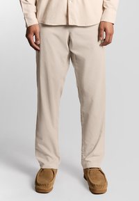 Mand iført beige bukser og en lys beige langærmet skjorte står med brune ruskind moccasin sko foran en ensfarvet baggrund.