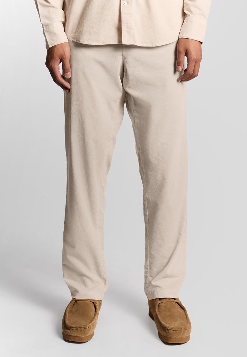 Mand iført beige bukser og en lys beige langærmet skjorte står med brune ruskind moccasin sko foran en ensfarvet baggrund.