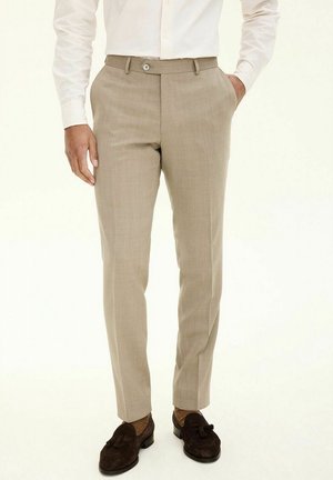Man draagt beige op maat gemaakte broek, wit overhemd met knopen, bruine loafers met kwastjes en geruite sokken, staat met één hand in de zak.