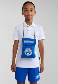 Blå tygaxelväska med en klaff, med en vit logo och text, bärs runt halsen, ihop med en vit polo och blå shorts.