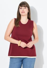 Vrouw met lang bruin haar in een mouwloze maroon top en lichtblauwe spijkerbroek, staande tegen een witte achtergrond met handen gevouwen.