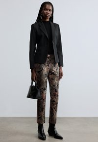 Blazer nero su misura sopra un maglione a coste nero, abbinato a pantaloni con motivo floreale in colori tenui. Borsa nera e stivaletti alla caviglia.