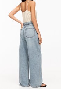 Mujer con pantalones de mezclilla anchos de tiro alto en un azul claro y blusa sin mangas beige, posando con una mano en la cadera, vista desde atrás.