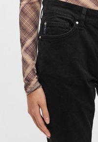 Jean noir en denim à coupe ajustée, avec des accents métalliques et une poche latérale. Main visible reposant sur la jambe du pantalon.