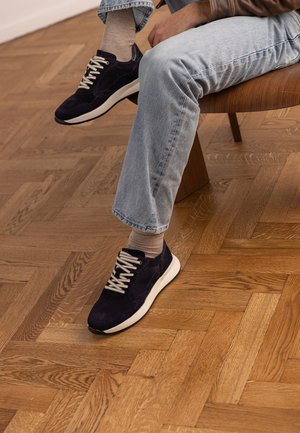 Person sitting on wooden chair wearing light blue jeans, beige socks, and black suede sneakers with white soles on wooden parquet floor.