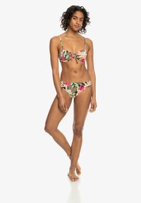 Femme debout pieds nus avec des cheveux bouclés portant un bikini à imprimé floral tropical avec des bretelles réglables sur fond blanc.