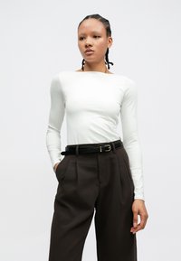 White long-sleeve shirt in a smooth fabric, paired with high-waisted dark brown trousers featuring a black belt with gold hardware detail.