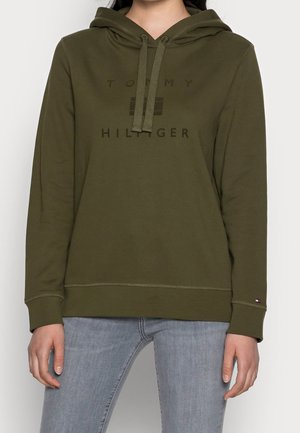 Kobieta w oliwkowo zielonej bluzie z kapturem Tommy Hilfiger z troczkami i jasnoniebieskich dżinsach, stojąca na białym tle.