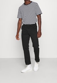 Pantalon noir fuselé à texture lisse, associé à une chemise à manches courtes rayée bleu marine et blanche. Porté avec des baskets blanches.