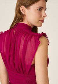 Mujer con ojos verdes que lleva una blusa sin mangas, transparente de color magenta, con detalles de volantes y pequeños botones a lo largo del cuello trasero, mirando hacia la derecha.