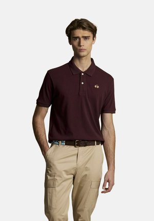 Giovane uomo che indossa una polo bordeaux scuro con logo ricamato e pantaloni cargo beige con le mani nelle tasche, in piedi davanti a uno sfondo semplice.