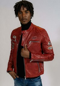 Veste de moto en cuir rouge avec des détails matelassés sur les épaules, des poches zippées et plusieurs patchs. Présente une coupe ajustée et une texture lisse.