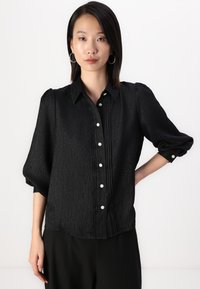 Black button-up shirt with textured fabric, featuring a collar, puffed sleeves, decorative white buttons, and a pleated front design.