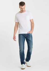 Camiseta blanca de cuello redondo con un bolsillo en el pecho, combinada con jeans azules. El conjunto se complementa con zapatillas blancas con suelas marrones.